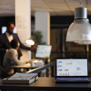 Laptop computer with financial graphs on screen, standing on table in startup office, coworkers discussing company strategy in background. Employees working late at night in startup office
